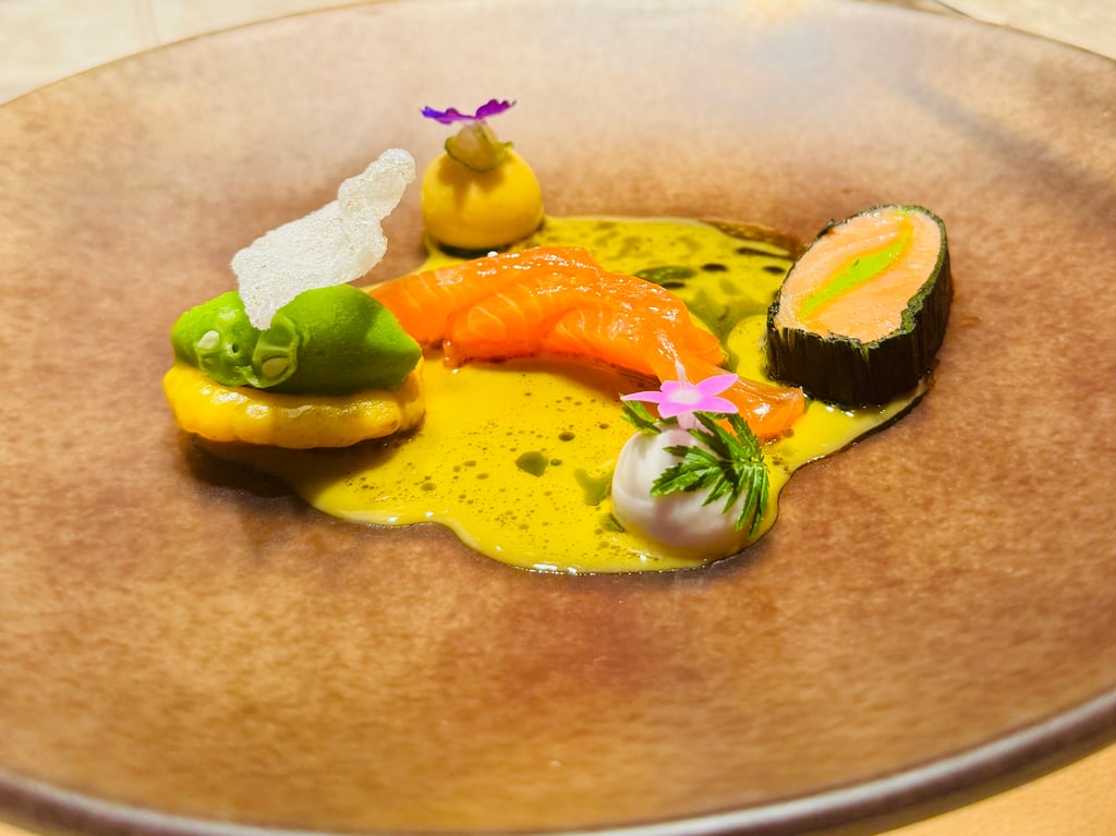 Assorted sushi on a yellow sauce: salmon nigiri, a seaweed-wrapped roll, green garnish, and a pink flower with decorative herbs.