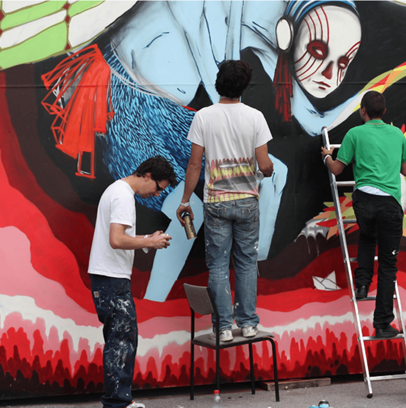 Three graffiti artists spray-paint a large mural on a wall: one standing on a chair, another on a ladder, and a third on the ground with a spray can, all amid a vibrant blue‑red artwork.