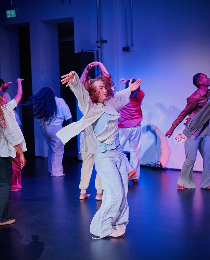 Dancers perform a contemporary routine in a blue-lit studio; foreground dancer raises arms while wearing light blue and white clothing.