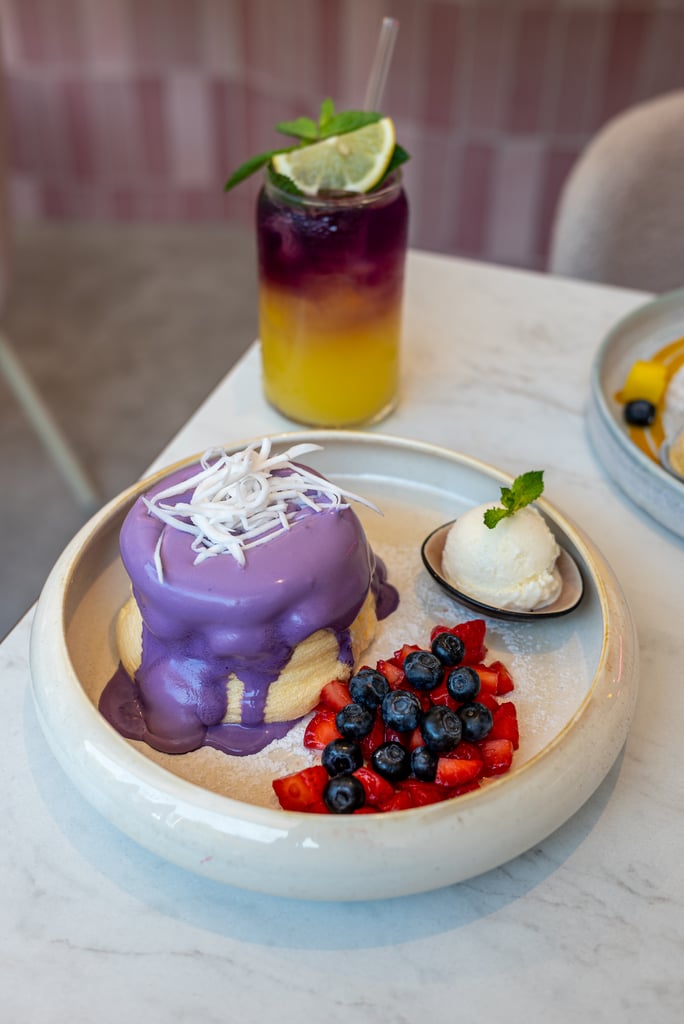 Plate with purple-glazed dessert topped with coconut shreds, mixed berries (strawberries and blueberries), and a small scoop of ice cream; mint garnish nearby on a white plate with powdered sugar. A layered yellow-to-purple drink with lime wedge sits in the background.