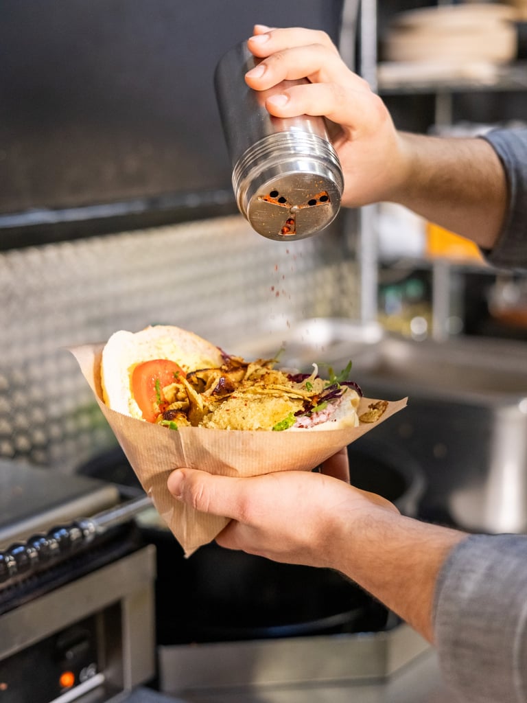 Person sprinkling red chili flakes from a shaker onto a wrapped sandwich held in hands in a kitchen setting