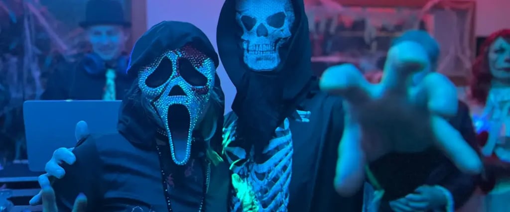 Two people in Halloween costumes with skull masks reach toward the camera under blue-purple lighting at a party.