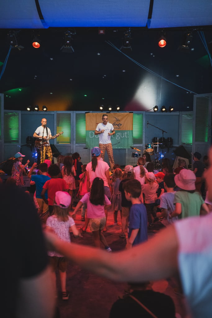 Children dance in front of a small stage as two musicians perform with guitars under colorful stage lights.