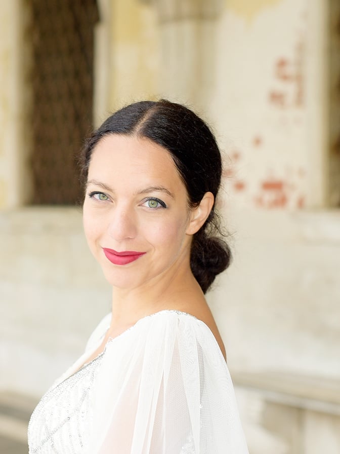 Woman with dark hair in a low bun, wearing a white dress, smiling softly outdoors in front of a light stone building.