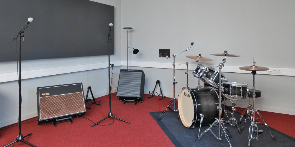 Drum kit with cymbals, two microphone stands, and guitar amplifiers in a practice room on red carpeted floor.