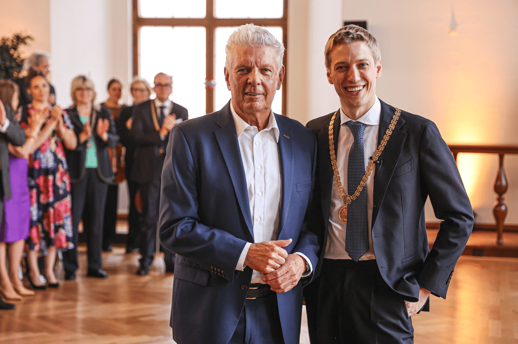 Two men in formal suits posing at an indoor ceremony; the man on the right wears a ceremonial chain while guests applaud in the background.