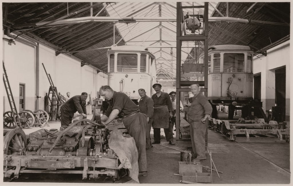 Inside a tram maintenance shop, workers repair a streetcar bogie at a workbench, with two trams in the background.