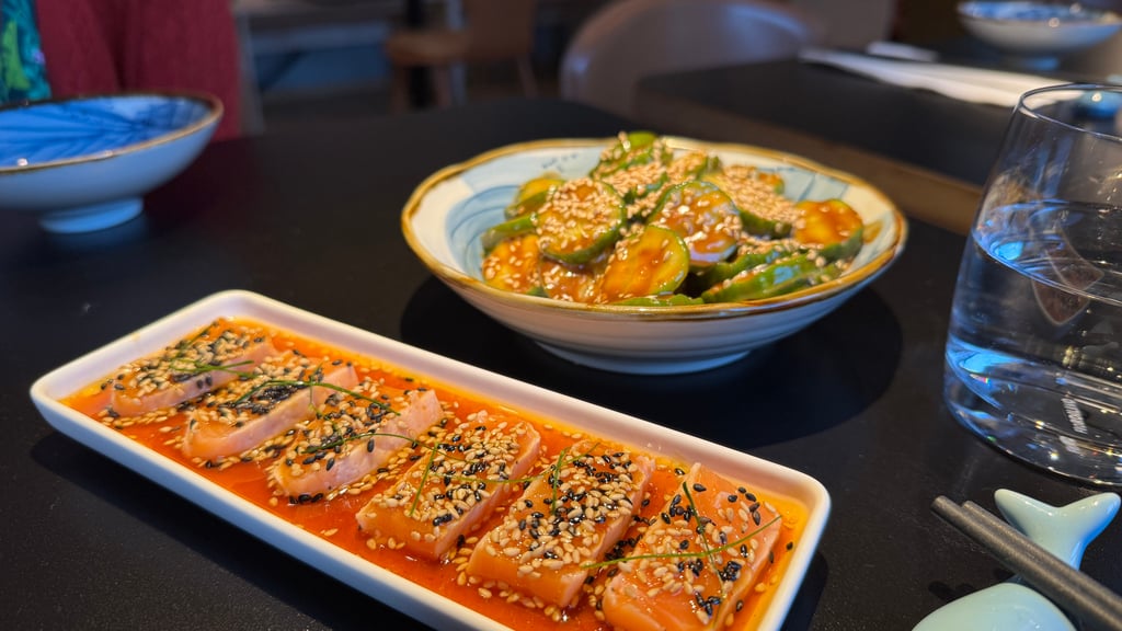 Salmon slices in orange sauce topped with sesame seeds on a white rectangular plate. A separate rectangular plate of sauce-glazed tuna or salmon with sesame appears nearby.