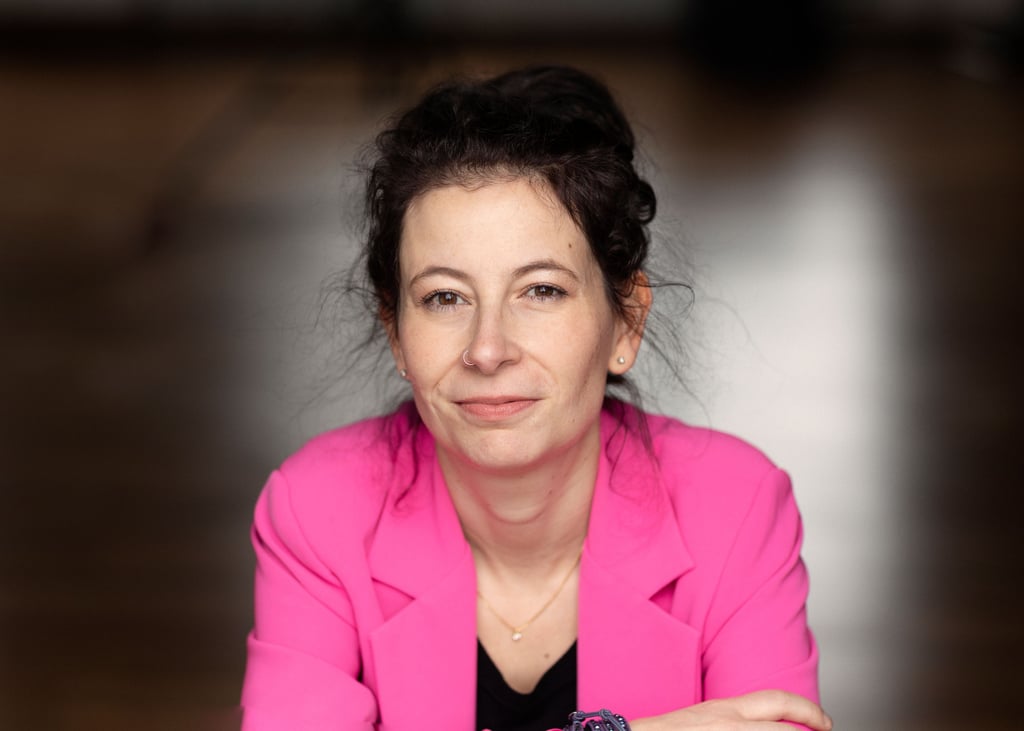 Close-up portrait of a woman with dark hair in a pink blazer, smiling slightly at the camera.