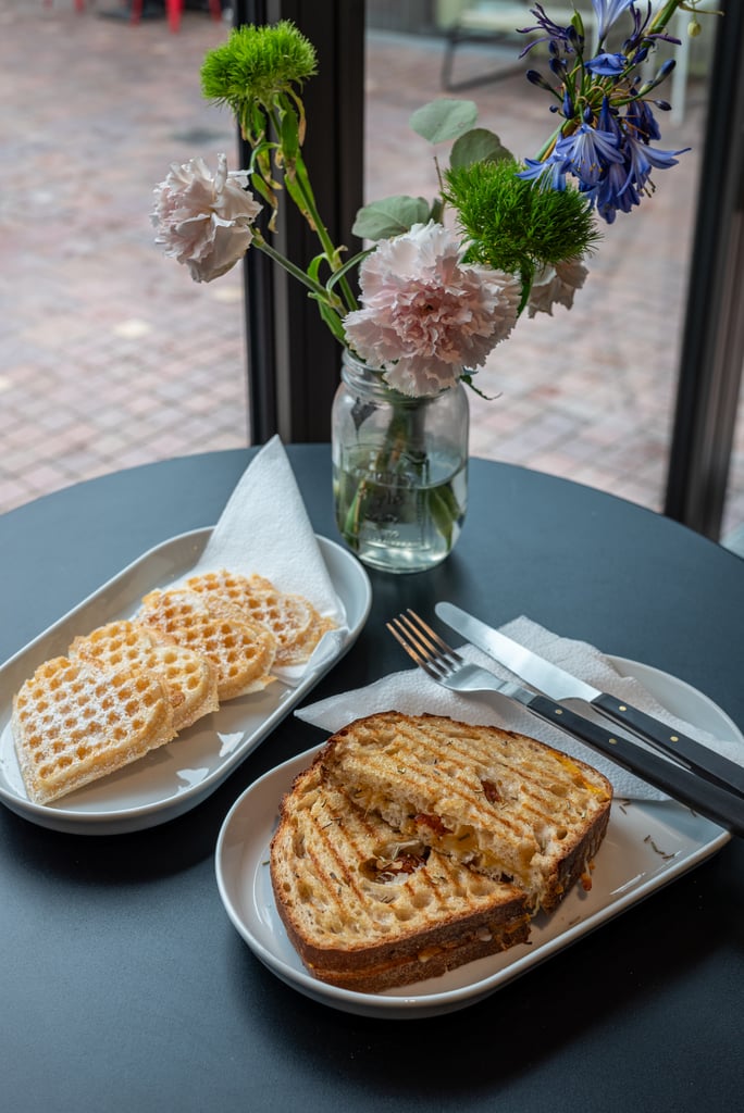 Waffle slices on a white plate with a grilled bread slice, set on a dark table by a window with a vase of flowers nearby.