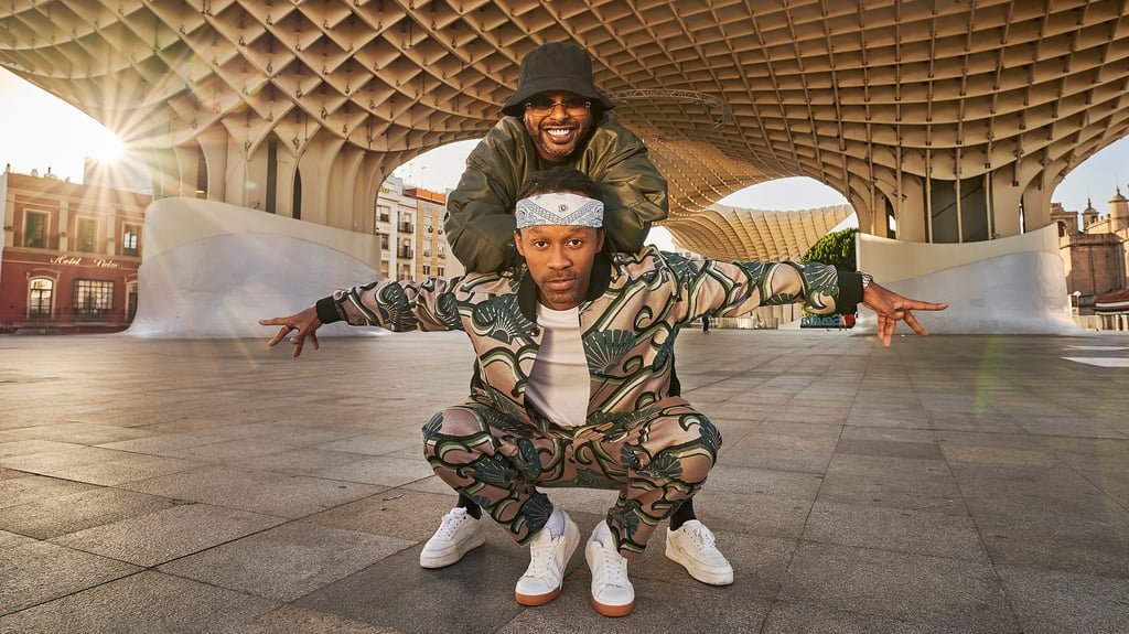 Two men perform a dynamic jump pose in an urban plaza under a curved, lattice-like canopy, sunlight streaming in from the left.