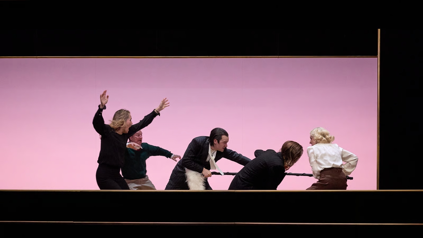 A group of actors on a pink stage, pulling a rope in a dramatic, playful tug-of-war scene.