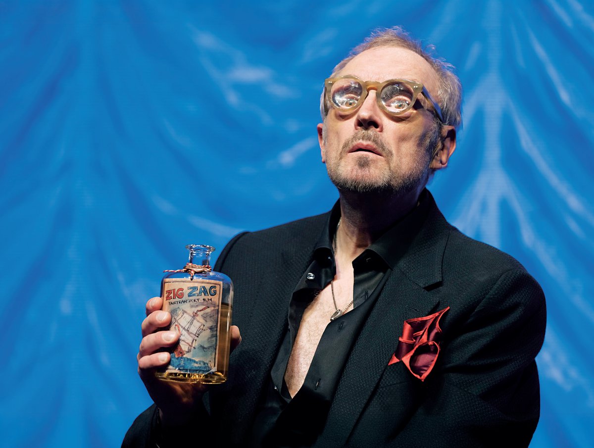 Man in a dark suit with a red pocket square holds a bottle labeled Zig Zag against a bright blue background.