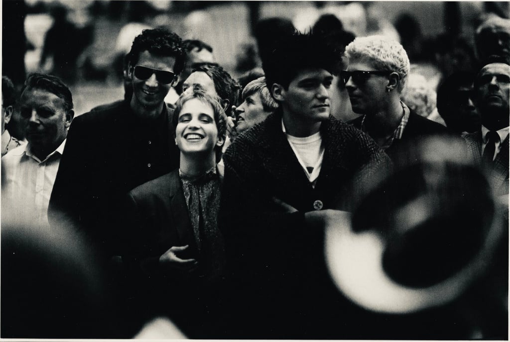 Crowd of smiling people at an outdoor event, with a woman at center laughing among friends wearing casual clothing and sunglasses in a black-and-white photo.