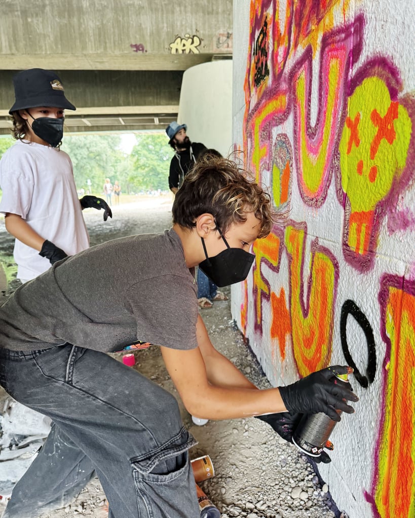 Person wearing a black mask and bucket hat spraying bright graffiti on a concrete wall under a bridge.