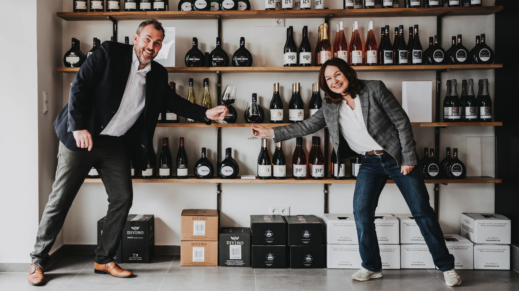 Two people in a wine shop clinking glasses for a toast with shelves of wine bottles in the background