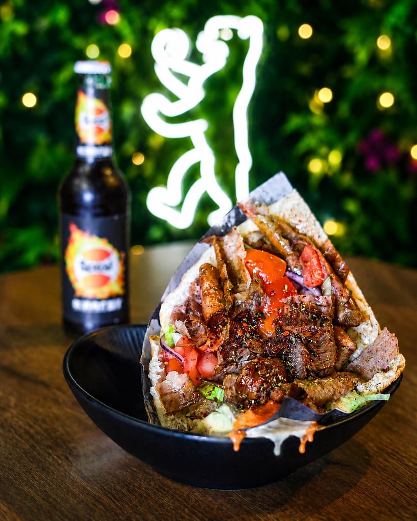 Giant taco stack with meat and vegetables in a black bowl; beer bottle and neon lights blurred in the background.