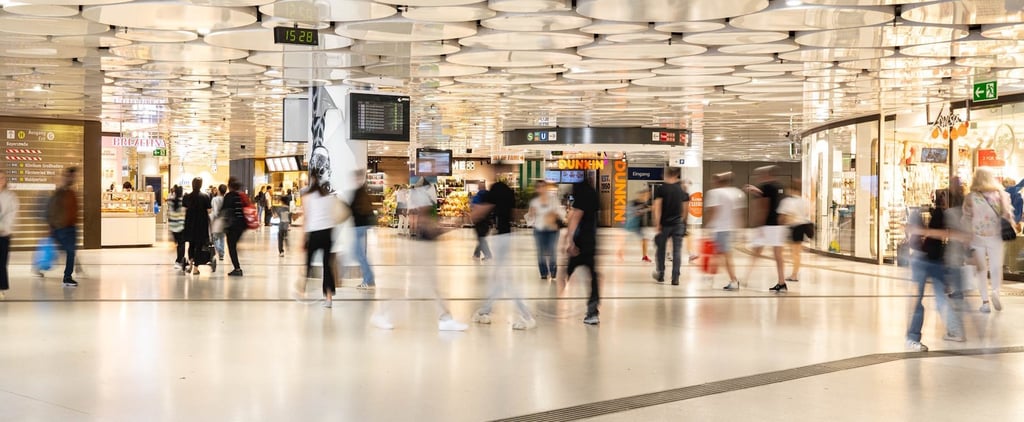 Busy indoor shopping concourse with blurred pedestrians walking past storefronts on both sides and circular ceiling lights above.