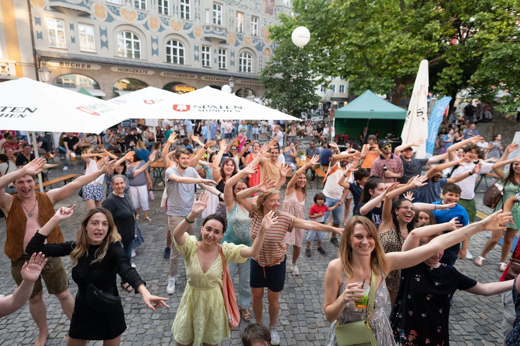 Crowd of smiling people in a city square raising their hands at an outdoor festival with tents and historic buildings behind them.