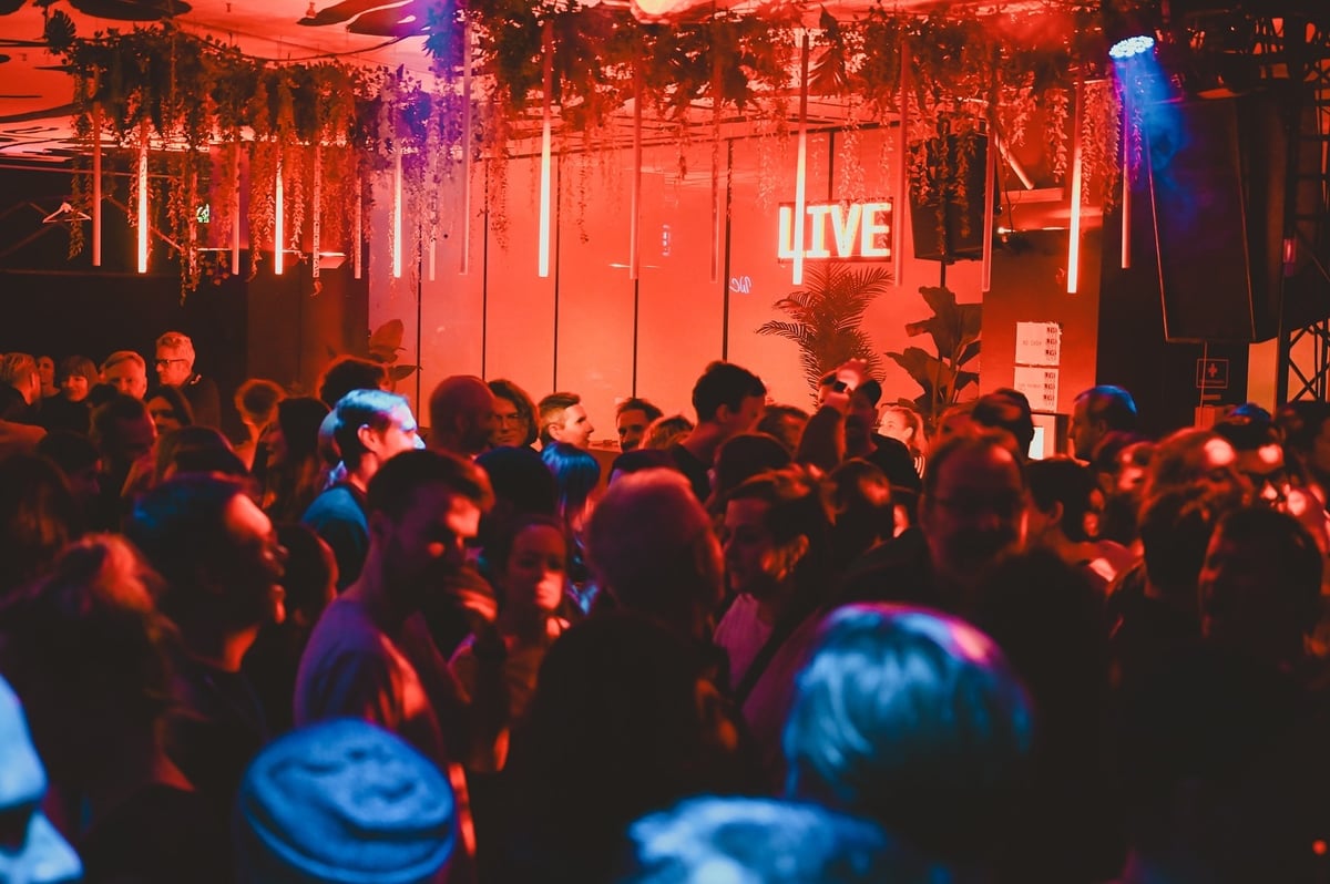 Crowded nightclub dance floor bathed in red and blue lights with hanging greenery and a neon LIVE sign in the background.