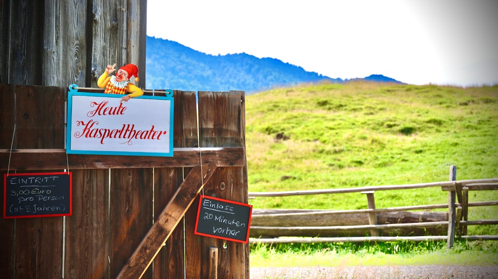 Wooden building exterior with a blue-framed sign reading 'Heute Kasperltheater' and a cartoon clown, set against a grassy hillside.