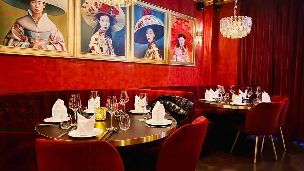 Elegant restaurant interior with red walls, gold-framed portraits of women in hats, crystal chandeliers, and round tables set with wine glasses and folded napkins.