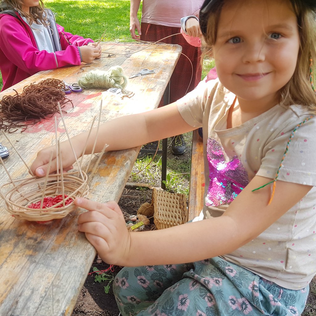 Smiling child in foreground at a wooden table weaving with thin sticks outdoors; several woven baskets and yarn on the table behind her.