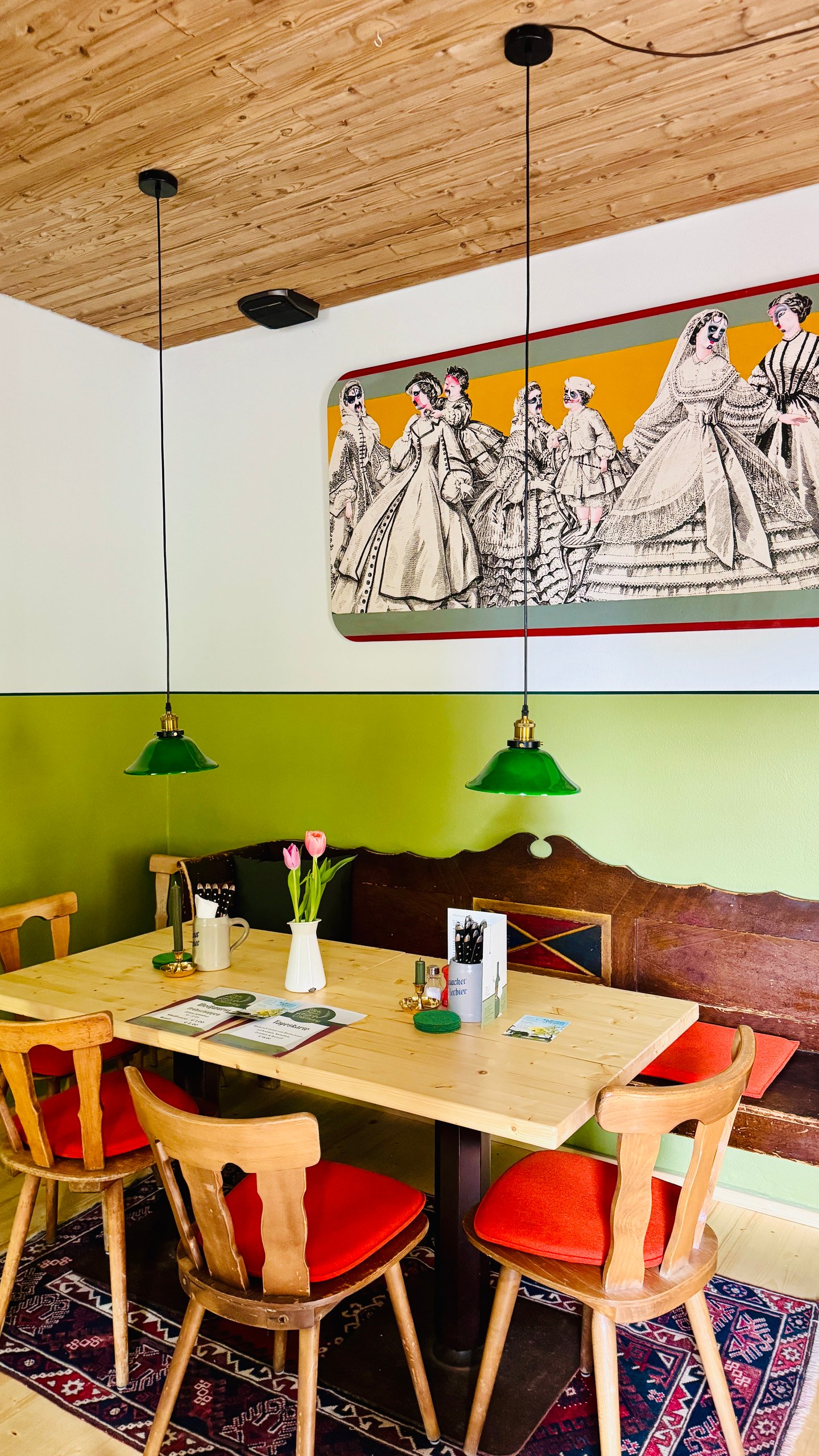 Cafe interior with a long wooden table, green pendant lights, a vintage mural, and wooden chairs with orange cushions.