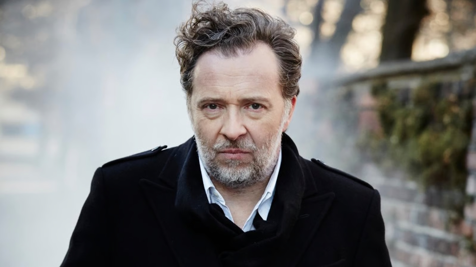 Portrait of a middle-aged man with salt-and-pepper hair and beard, wearing a black coat, looking at the camera outdoors.
