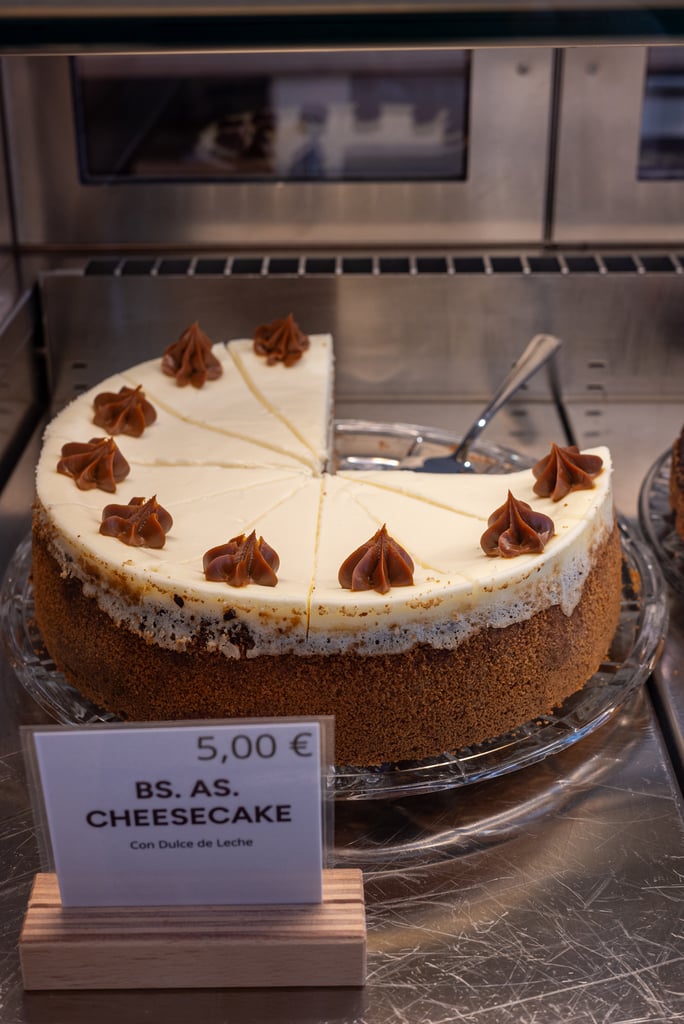 Whole cheesecake with white frosting and chocolate swirls on top, displayed in a bakery case (€5).