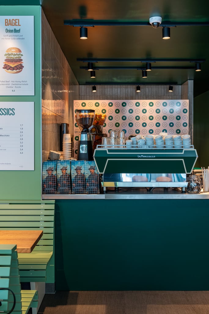 Cafe counter with an espresso machine, stacked cups, and a patterned wall; menu boards on the left.