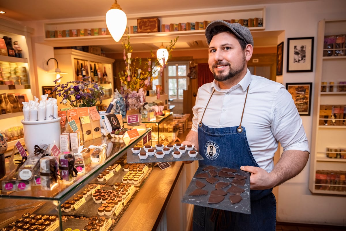 Smiling chocolatier in an apron holds a tray of assorted chocolates behind a glass display in a shop