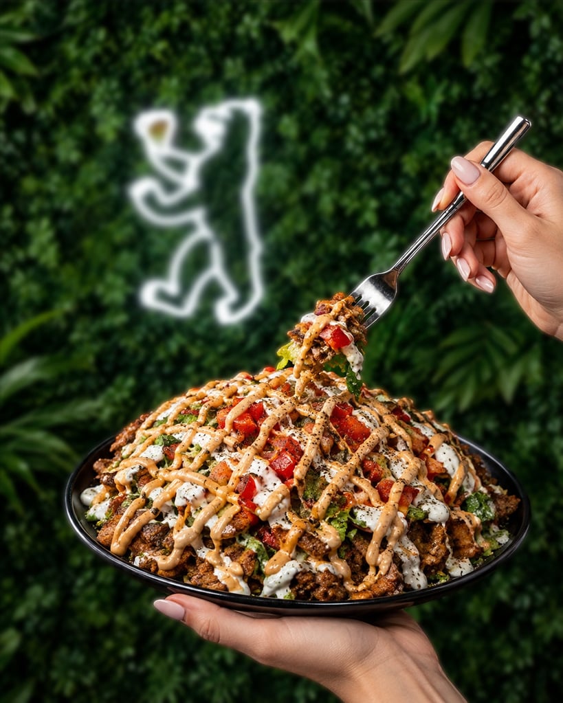 A hand holding a fork lifting a loaded plate of nachos drenched in crema and sauce, with vegetables on a dark bowl.