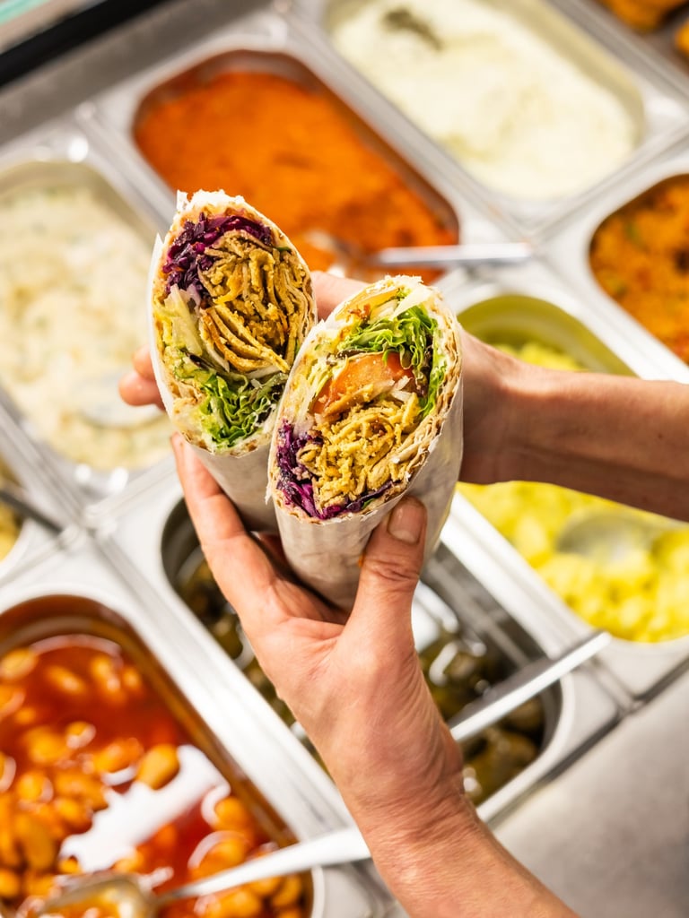Two halves of a wrap held by hands, filled with shredded chicken, lettuce, purple cabbage, and other veggies at a cafeteria buffet behind them.