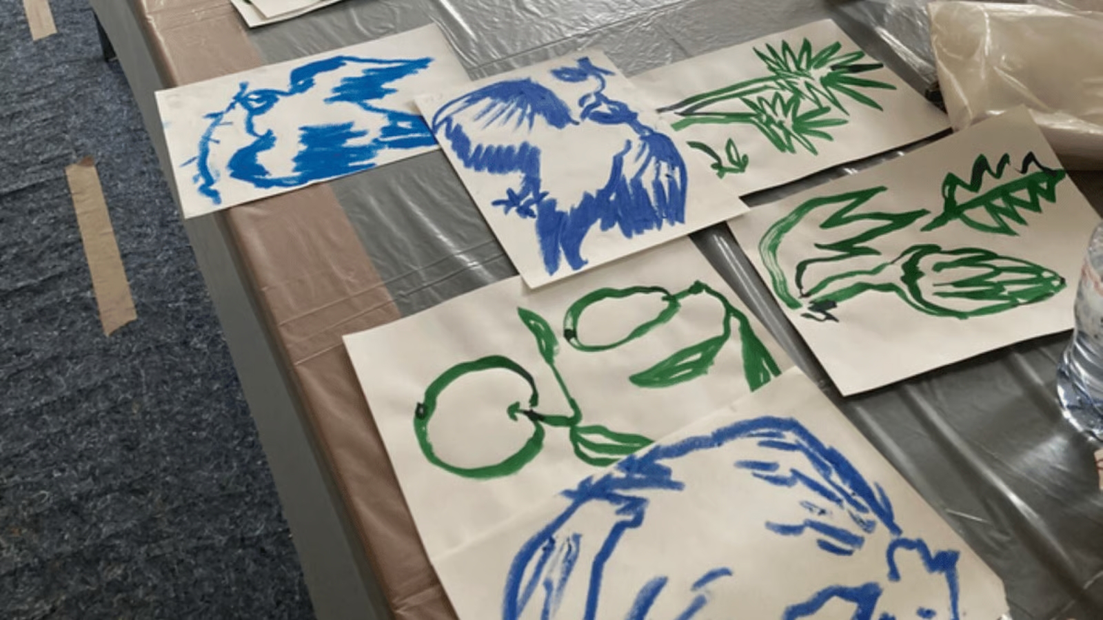 Several sheets of white paper with blue and green abstract brush drawings of fruits and plants spread on a plastic-covered table.