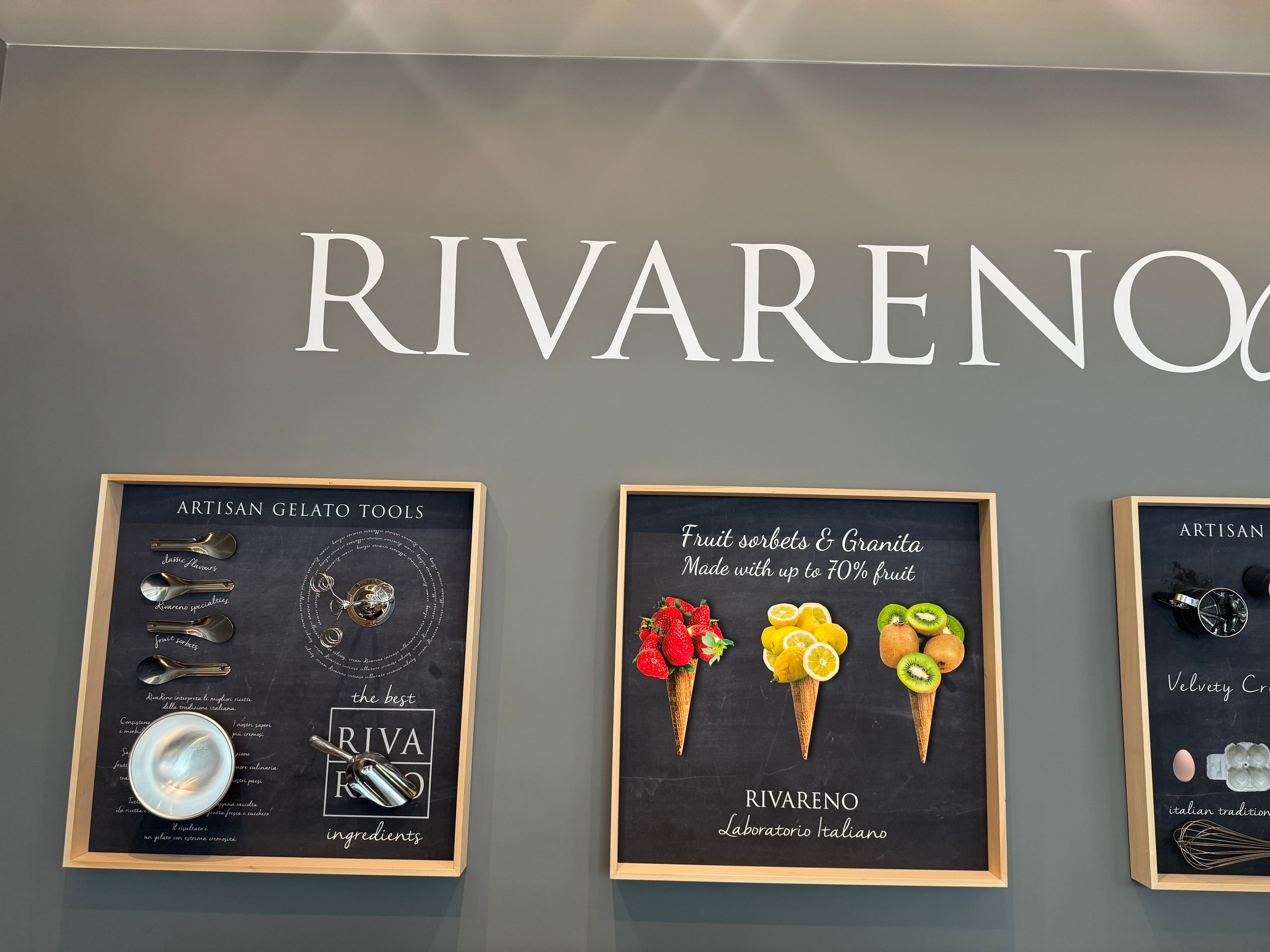 Wand in der Eisdiele RivaReno mit dem Schriftzug und drei beleuchteten Tafeln zu Gelato-Werkzeugen und Fruchtsorbets
