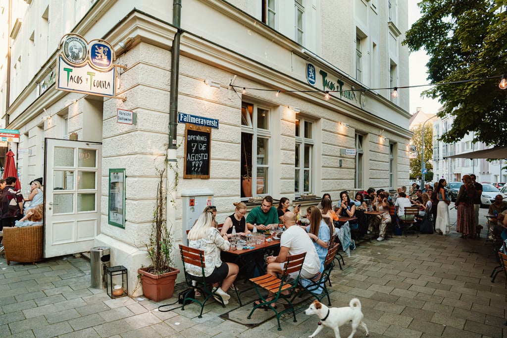 Outdoor restaurant on a sunny street; diners fill long tables outside a white building, with string lights overhead and a white dog in the foreground.