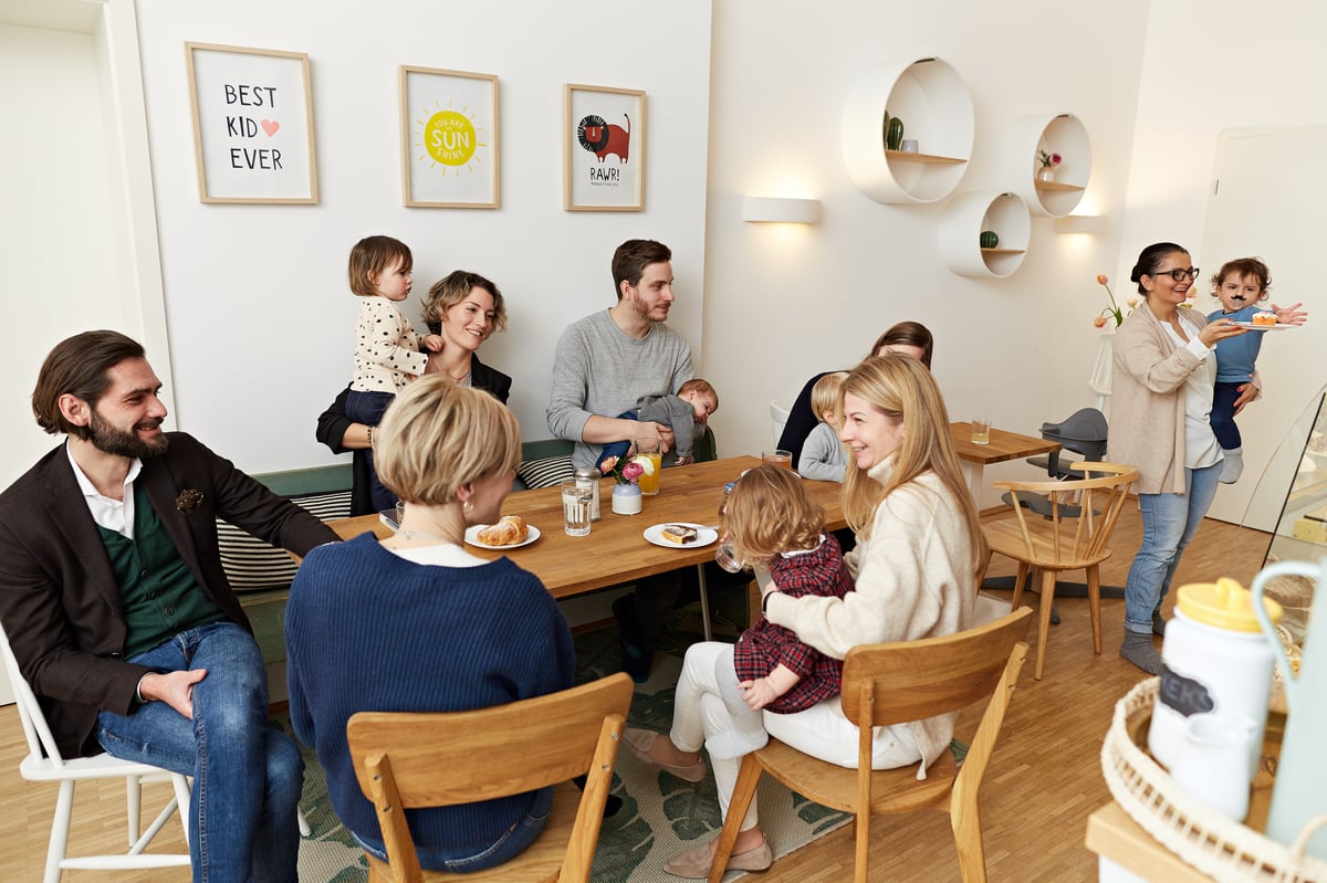 Familien mit Kleinkindern und Babys sitzen im Café um einen Tisch herum und trinken Kaffee und essen Kuchen