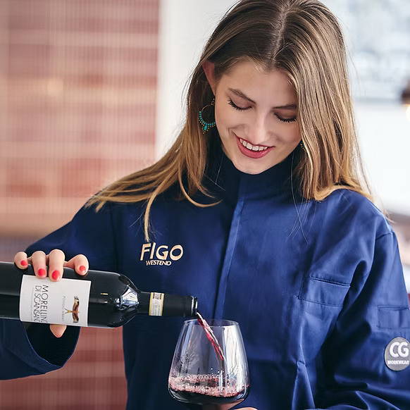 Woman in a blue jacket pours red wine into a glass and smiles.