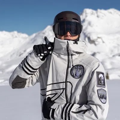 Snowboarder in a white jacket and black goggles makes a peace sign in a snowy mountain landscape.