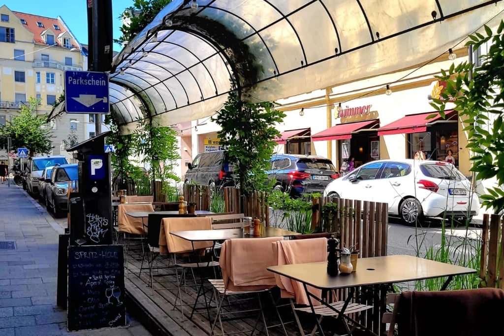 Outdoor city café terrace with beige tables and peach-colored chairs under a curved, translucent canopy, parked cars lining the street behind a wooden fence.