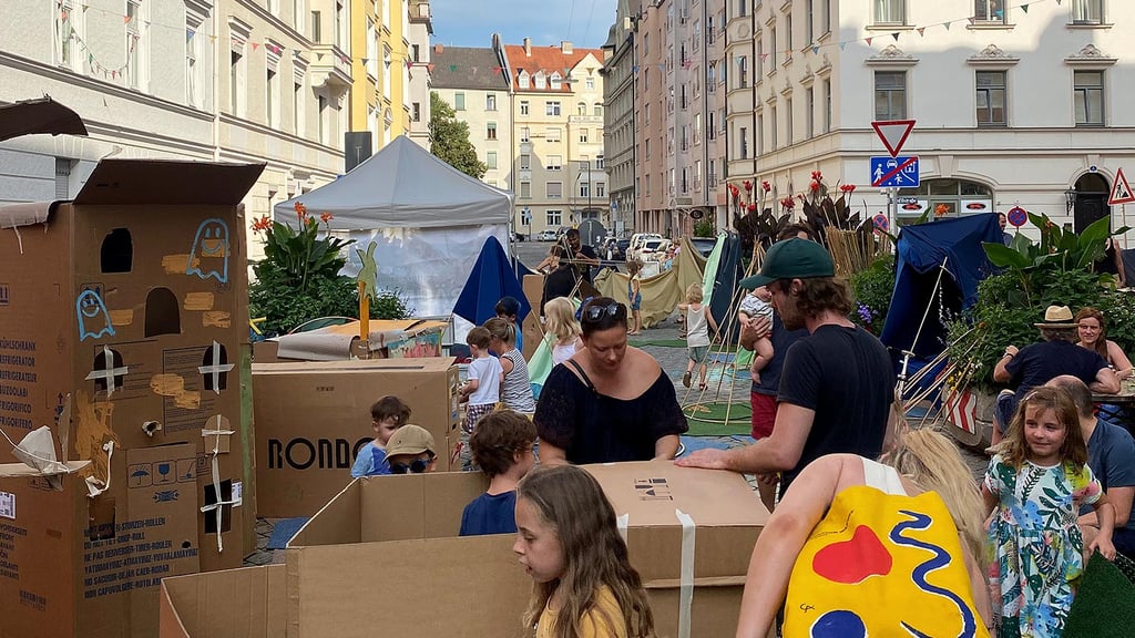 Kinder und Erwachsene bauen in einer autofreien Straße mit Kartons und Zelten fantasievolle Spielhäuser beim Straßenkunstprojekt Straße. Oase in München