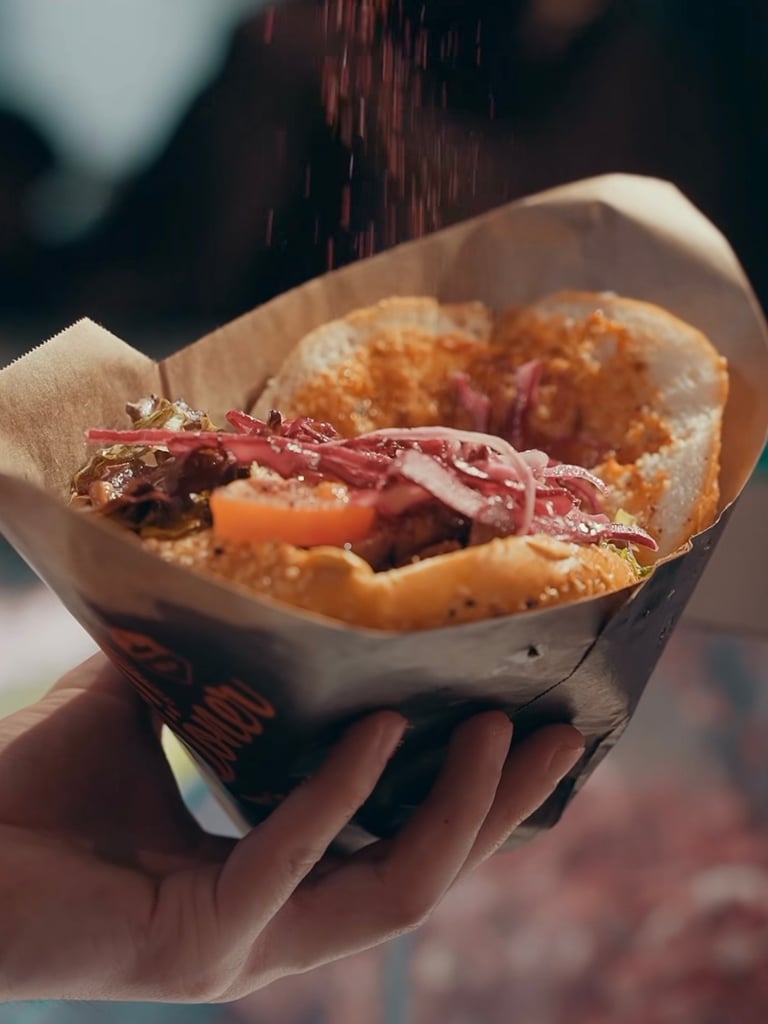 Hand holding a loaded sandwich in brown paper wrap, topped with red onions and greens.