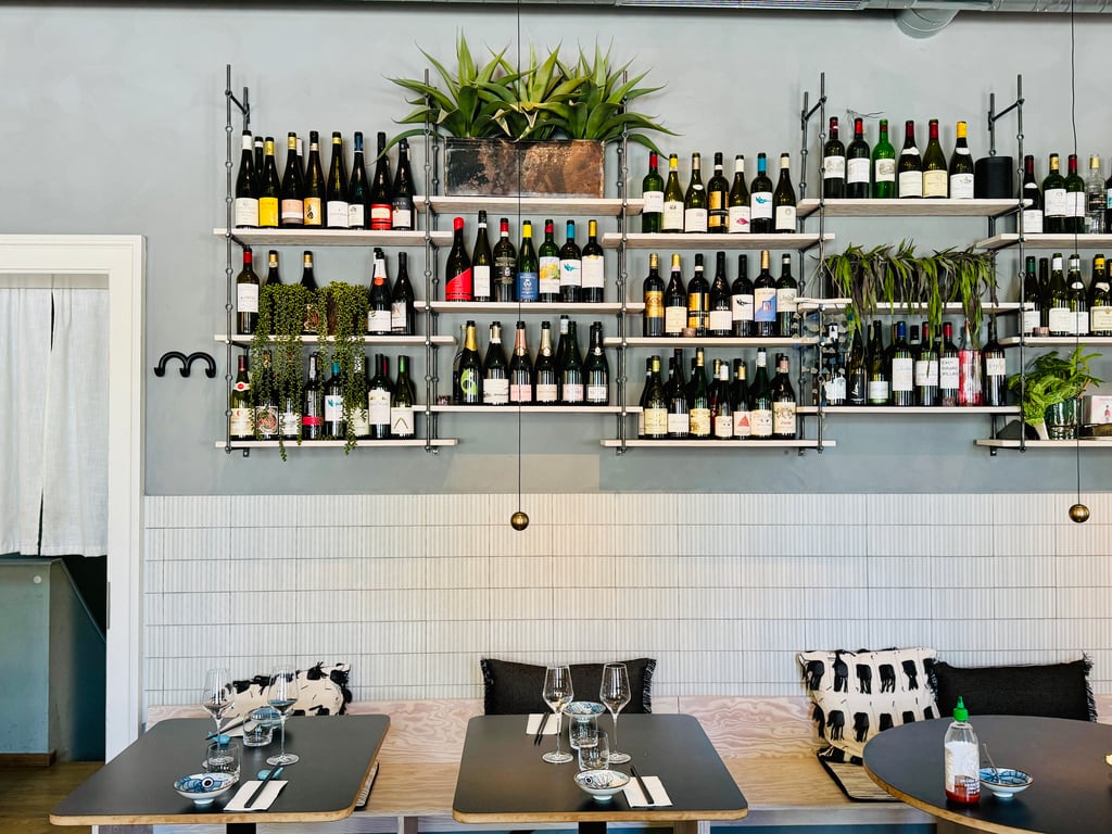 Wine bottles arranged on industrial shelves behind a dining table with glasses and plants in a modern restaurant interior.