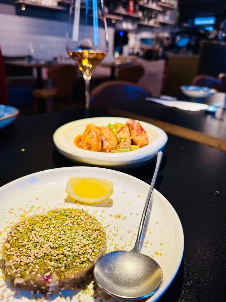 Plate with a sesame-seed crusted dish and a lemon wedge, spoon nearby; blurred wine glass and restaurant background.