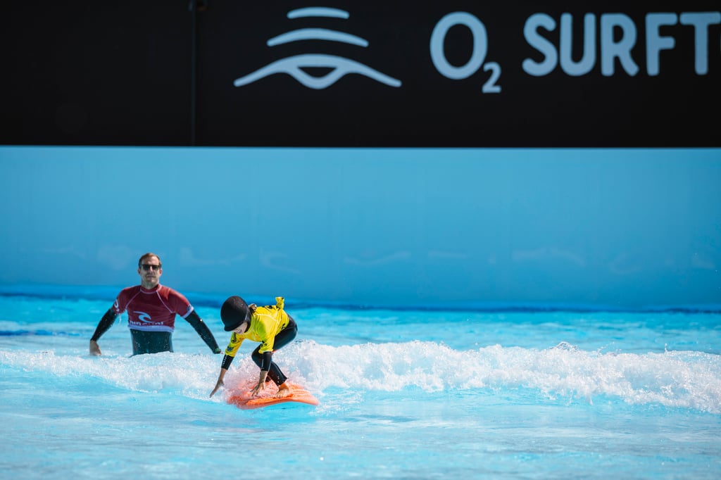 Two surfers ride small waves in bright blue water, with a dark wall displaying light-blue 'O2 SURF' text in the background.
