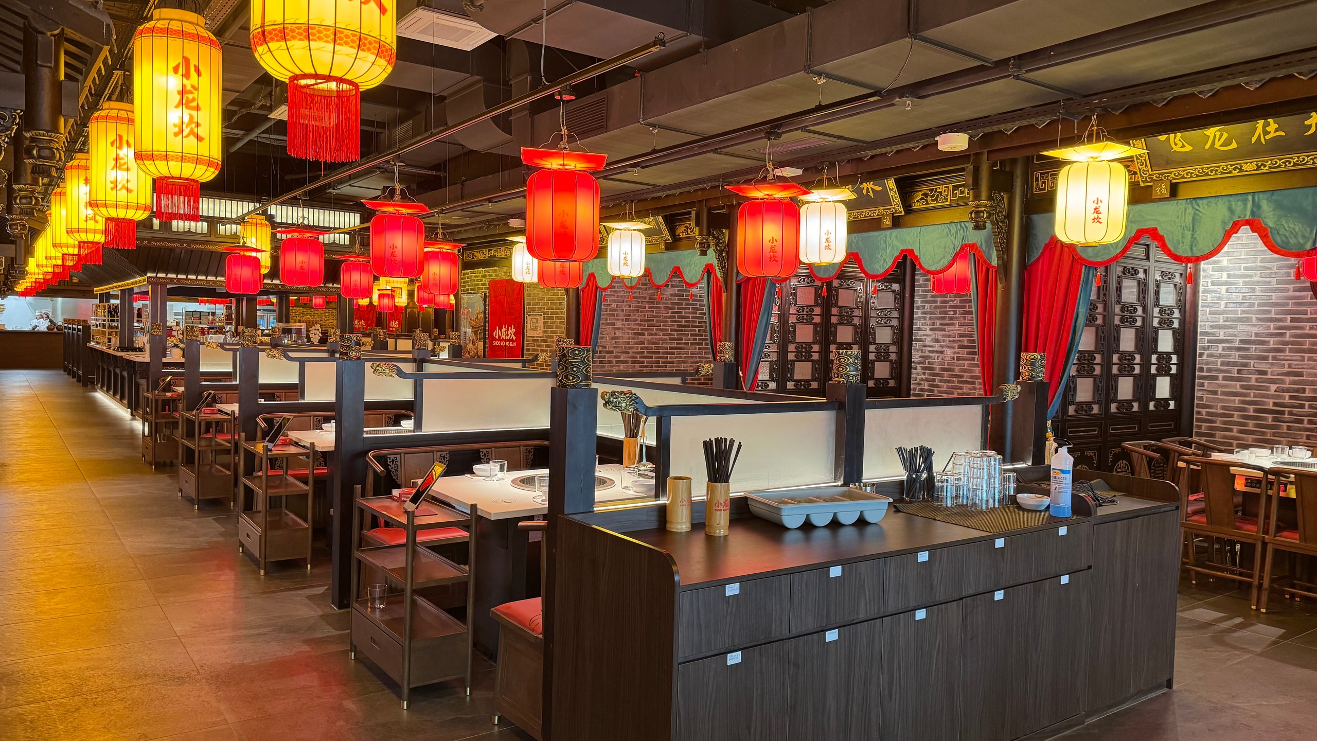 Interior of a Chinese restaurant with red and yellow hanging lanterns, dark wood booths, brick walls, and tables set for dining.