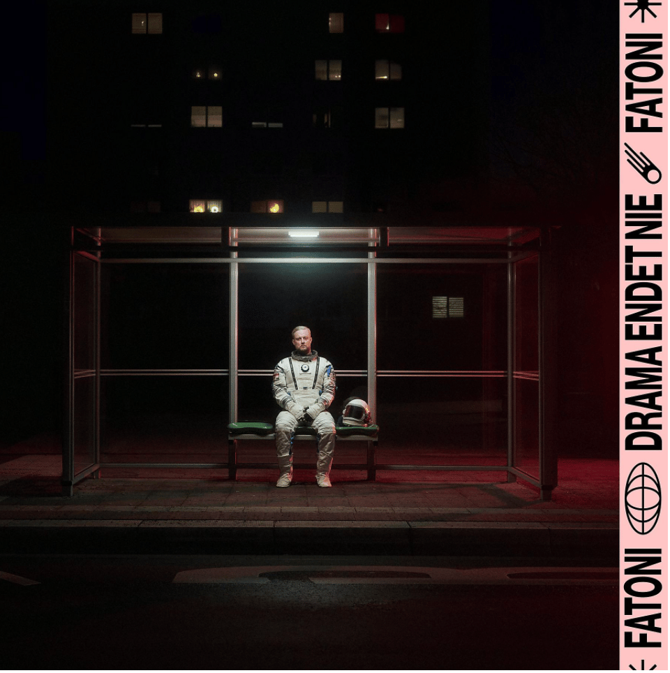 A man in a white space suit sits alone on a green bench inside a glass bus shelter at night, with city lights behind him and a pink vertical banner along the edge.