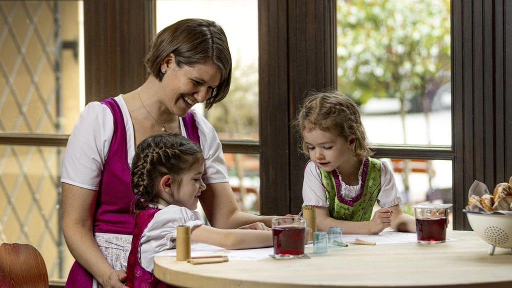 Mother helping two young girls with a craft activity at a round table, smiling warmly at them.
