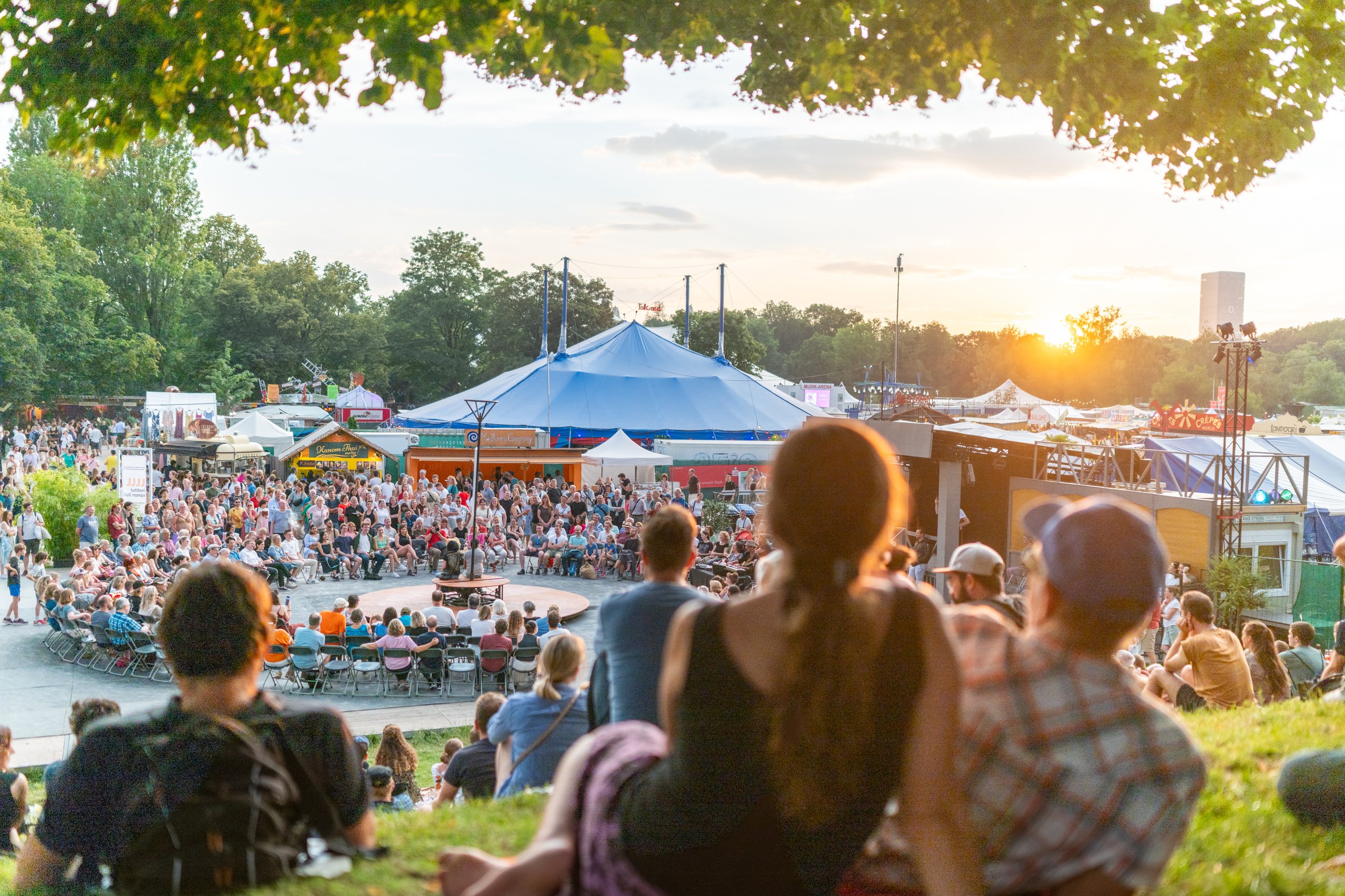Crowd seated around an outdoor festival stage with a large blue tent and food stalls at sunset.